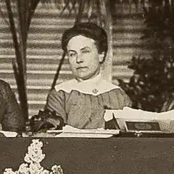 A woman in a dark dress with a wide white collar sitting behind a dais which has papers on it and horizontal striped panels and plants in back of her.
