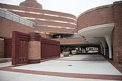 The entrance to the Administration Building, which consists of an open-air walkway running under a brick building with a rounded facade. In the foreground is a red gate, while on the right, part of the building overhangs the walkway.