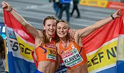 Photo of Femke Bol and Lieke Klaver posing for the camera, while holding Dutch flags with the texts 'Giro 555' and 'Geef nu' meaning 'Donate now [to] giro [account number] 555'
