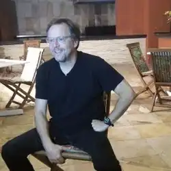 A color photograph of a man sitting on a sofa, with two golden statuettes on the table in front of him.