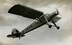 Black-and-white photo of a small propeller airplane, seen from below while climbing. Its landing gear is long and not foldable. It sports a Nazi Germany tail decoration and the sign D-IKVN.