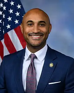 Official House portrait of Figures smiling in front of the U.S. flag, wearing a dark blue suit, white shirt, and checkered red and gray tie.