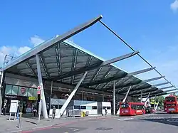 A large modern building with a massive canopy extending upwards from the top and over the bus station, which is supported by large V shaped pillars. An underground roundel is visible behind one of the windows and two buses are parked in the station.