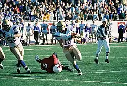 Several men dressed in football uniforms dart down a field. Another football-suited man is collapsed on the ground, while a man is scene in the background wearing a black and white striped shirt holding a whistle. Behind the scene are hundreds of people cheering in stands.