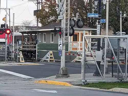 A beige Craftsman station house, situated at the right of two tracks, with a red peaked roof and a double beige door.