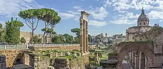 The image shows the ruins of the Forum of Caesar in Rome. Three tall ancient columns dominate the center, surrounded by scattered stones and fragments of walls covered in patches of grass. On the right, the dome of a church rises above the ruins. Tall pine trees stand to the left, and the background features modern buildings. The sky is bright and partly cloudy.