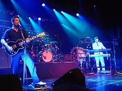 A three-piece guitar playing a live gig. Morgan stands at the front of the stage, playing guitar and with his mouth to a microphone on a stand.