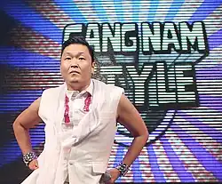 A picture of Psy wearing a white vest, posing in front a multicolored background with a sign reading "Gangnam Style".
