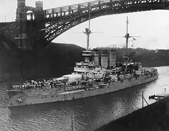 A large warship, bristling with guns, passes under a high arch bridge through a narrow canal.