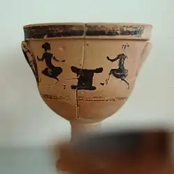 A small, ceramic, cream to light brown colored cup with a painting in black depicting two girls dancing around a shrine to Artemis
