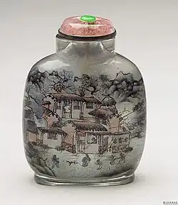 An inside-painted snuff bottle with a stout top and a short tan stopper topped with a green finial. It features a scene of houses in a forested rural environment