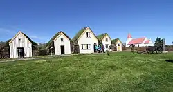 Several turf houses in a row with a broad field of grass in front of them. There is a church a little ways from the houses.