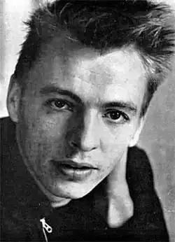 Black and white close up of a slim white man with short hair at the sides and piled up on top like a brush wearing a black shirt