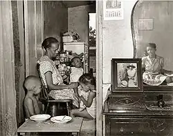 Black and white photo of a room; on the left side is a doorway through which Watson and her children can be seen sitting in a room. On the right side is a drawer, upon which rests a mirror reflecting a different woman and a framed photoportrait of two people.