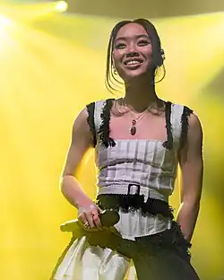 Griff wearing a white dress with black details on stage. She is smiling and holding a microphone in her right hand.