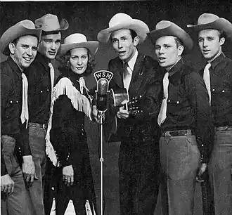 A group of six men and one woman wearing cowboy hats, standing around a microphone. The man third from right is holding a guitar.