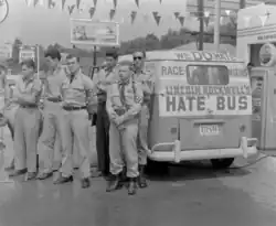 Black and white photo of Burros standing with several other men wearing swastika armbands in front of a band labeled "Lincoln Rockwell's Hate Bus" and "We Do Hate Race Mixing"