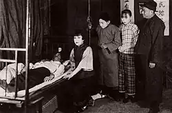 A black-and-white image of a young woman kneeling beside a bedridden man, with three people standing to her left