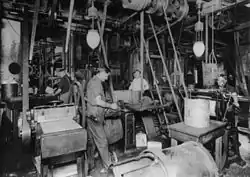 Photograph of a large workshop crowded with machinery. There are at least four men working there. There are belts coming down from the ceiling that drive the machinery. Two electric lighting fixtures are also hung from the ceiling.
