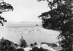 A view of yachts in a bay, with wooden buildings on the beach. A small island is visible in the distance.
