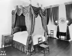 A black-and-white image of one of the bedrooms as it appeared in 1936. There is a four-poster bed in the center of the room, a cabinet to the left, and chairs in front and to the right. The wall on the right has some curtains.