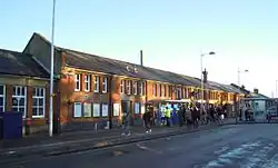 A very long red-brick two-storey building with a slanted roof and chimneys as photographed from across the street. A large number of people block the view of the ground floor of the building.
