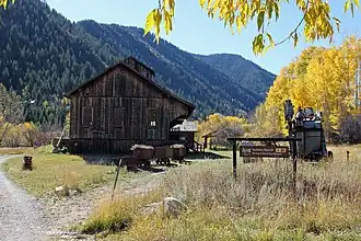 A wooden building with a pointed roof between two dirt paths, with some machinery next to it on the right. In the rear left are wooded mountains, and a stand of trees with yellow leaves is on the rear right.