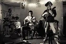 A three-piece band, consisting of drums, acoustic guitar, and vocals/ukulele playing in a home. The ukulele player/singer is wearing a pirate hat