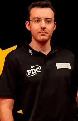 Headshot of Huw Ware referending for the Professional Darts Corporation. He is a white man with dark hair and beard, wearing square glasses.