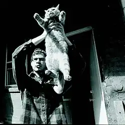 A black-and-white image of a man smoking a cigarette in a flannel shirt and a backwards baseball hat holding up a cat by its armpits