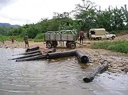 Eight rosewood logs lying at the riverside with nearby workers, a truck, and a cart