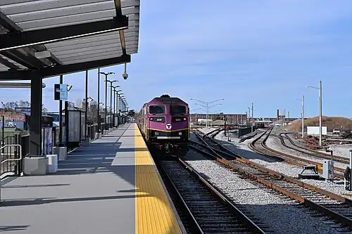 A diesel locomotive trailing a passenger train leaving a station with a high-level platform