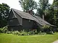 A large brown colonial-style house with multiple leanto additions. There is a flower garden in front of the house.