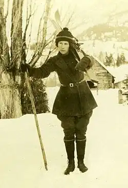 A sepia toned photograph of a dark haired woman in winter clothing holding skis on her back with her left arm and a hiking stick in her right hand. The background is snowy and mountainous, featuring a bare tree and a cabin.