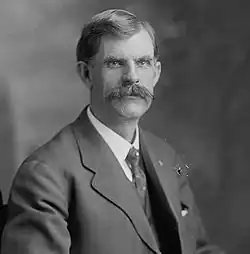 A man with dark hair and a mustache wearing a dark jacket, patterned tie, and white shirt
