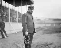 A greyscale photograph of a baseball umpire standing near the bleachers, he is wearing a suit and newsboy cap, and is holding his mask in his left hand.