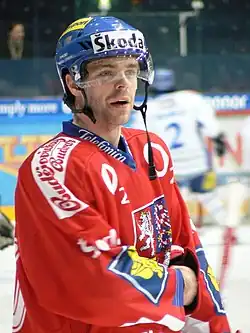 An ice hockey player standing, looking to the right of the camera. He is wearing a blue helmet and a red uniform.