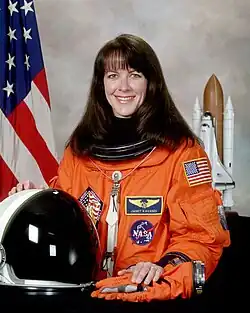 image of Janet Kavandi in 2001, posing in her orange NASA uniform with helmet in front of her, and U.S. flag and an upright Space Shuttle model to each side of her in the background
