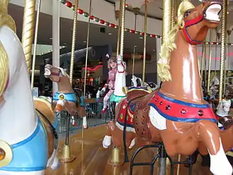 A close-up of a brown carousel horse with a brown saddle and a red bridle on a historic carousel with a girl riding a white carousel horse in the background