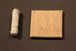 A carved cylindrical object and a small plaque of clay showing a repetitive geometric design