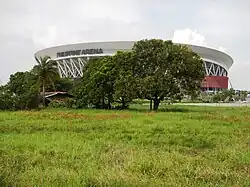 A large stadium with the name "Philippine Arena" displayed on its facade, surrounded by lush green grass and several trees in the foreground. The sky is partly cloudy.