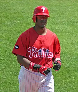 A dark-skinned man in a red baseball jersey and red left-handed batting helmet walks on a baseball field; he appears to be in his mid-twenties. His jersey reads "Phillies" in white and red script, with two blue starts dotting the "i"s.