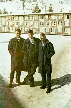 Black and white photo of three men wearing full-length suitcoats, standing in front of a dormitory building
