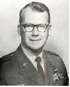 Black and white portrait of a caucasian male, short dark hair, dark rimmed glasses, clean shaven; wearing a light colored shirt, dark tie, dark jacket. Jacket has US emblem on left lapel; military ribbons on right chest