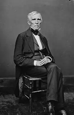 A man with gray hair wearing a black suit and white shirt, sitting with his hands folded