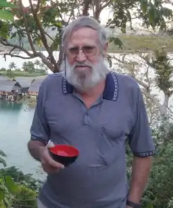 An elderly man in a loose blue polo shirt with a massive beard and huge aviator glasses holding a small black bowl which is red on the inside; the background is tropical with indigenous housing in the distance