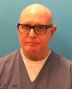 A mugshot of a clean-shaven bald white man; wearing a grey-and-white prison uniform and black spectacles, he is facing and looking into the camera.