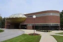 View of the theater's facade as seen from a nearby sidewalk. The sidewalk curves left in the background, with two paths splitting off to the right and a curving road to the left. Behind the sidewalk is the theater, which is flanked by brick pavilions.