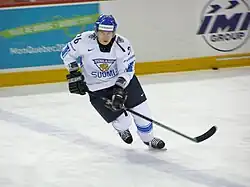 An ice hockey player standing directly in front of the camera. He is wearing a black helmet and uniform.
