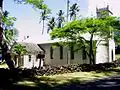 Photograph of the church established by Father Damien at the Kalaupapa Leprosy Settlement, behind a low stone wall and surrounded by palm and other trees.